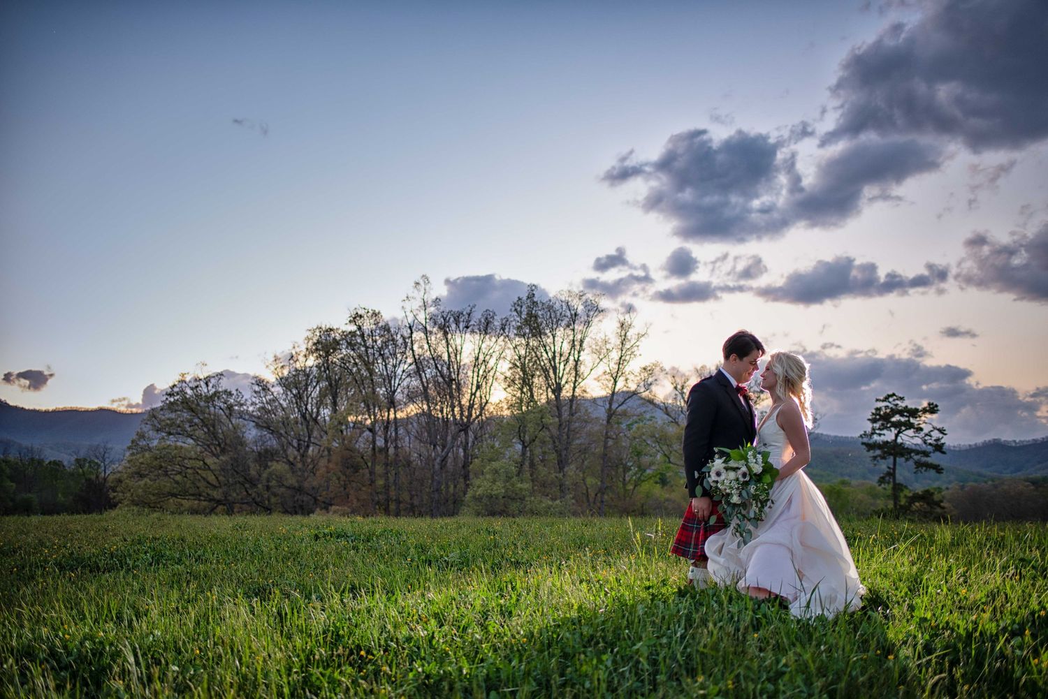 Bride and groom share a romantic kiss in a lush green field at sunset with mountains in the background.