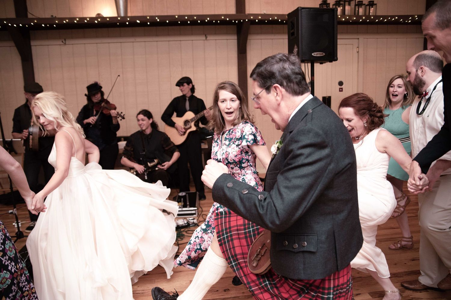 Lively wedding reception dance floor with guests in kilts and formal wear, band performing in background.
