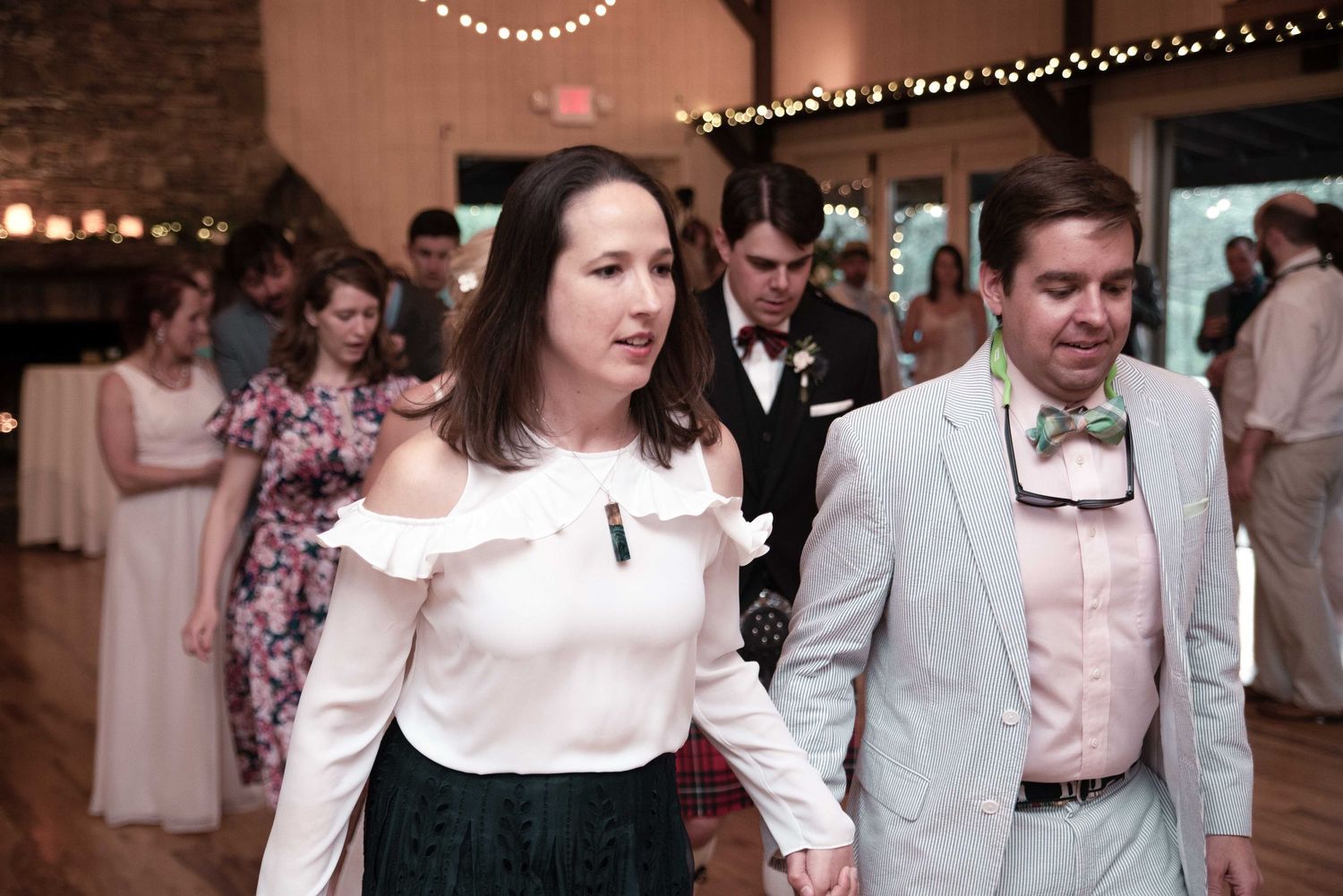 Couple holding hands on a wedding reception dance floor, woman in white off-shoulder top, man in seersucker suit with bow tie.