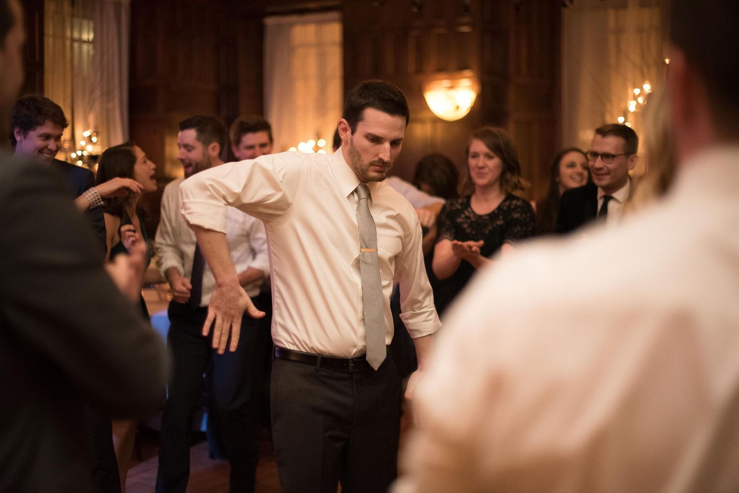 Man dancing energetically at a wedding reception surrounded by guests in formal attire in an elegant venue.