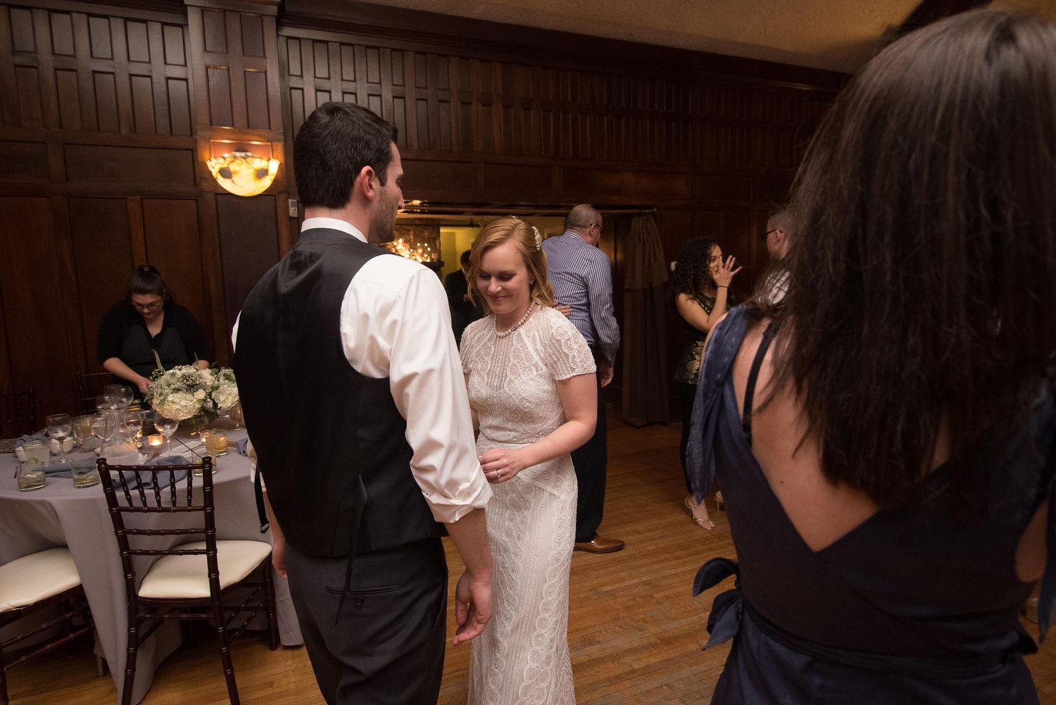 Bride and groom share a dance at elegant wood-paneled wedding reception venue with guests in background.