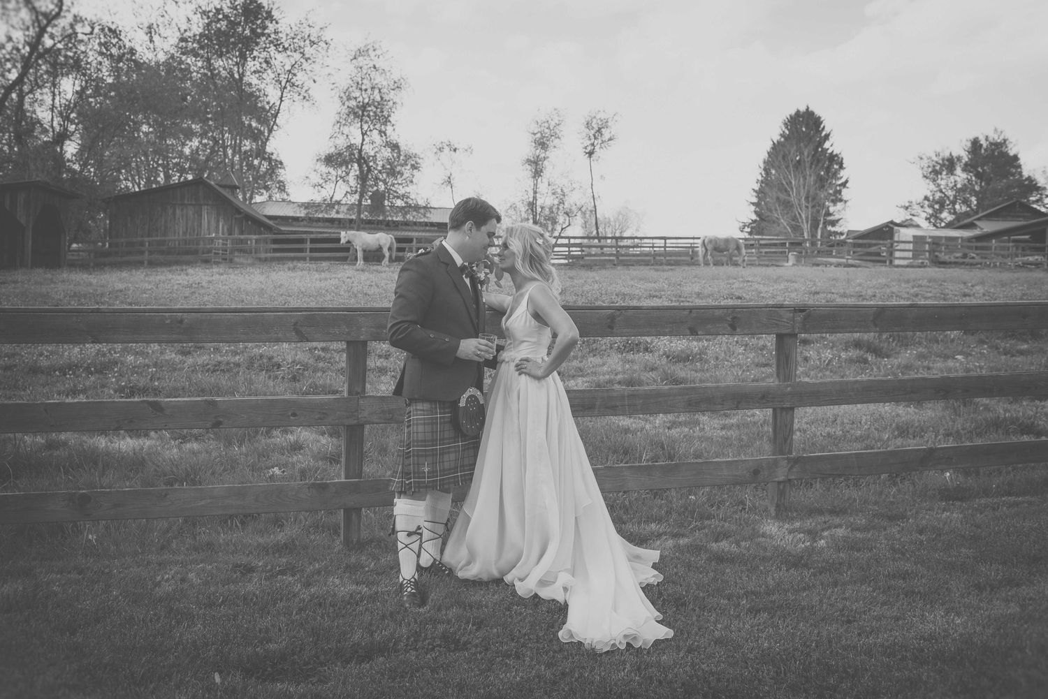 Black and white photo of a bride and groom sharing a romantic moment by a wooden fence at a rustic outdoor wedding.