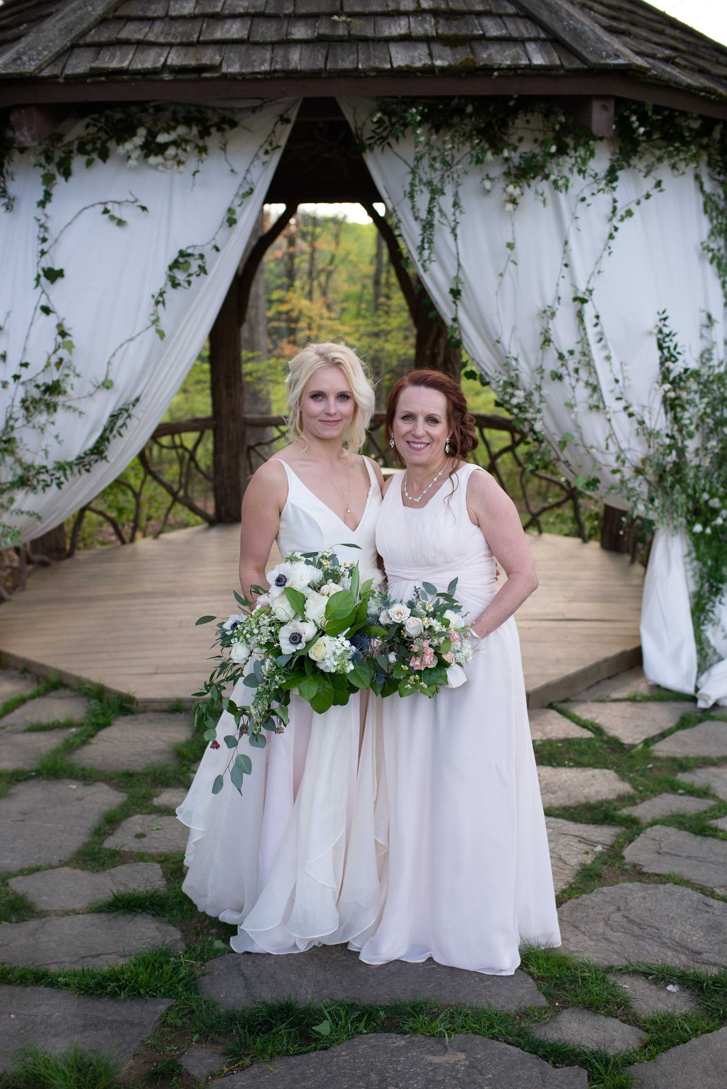 Two brides in white gowns holding bouquets stand before a floral gazebo on their wedding day.