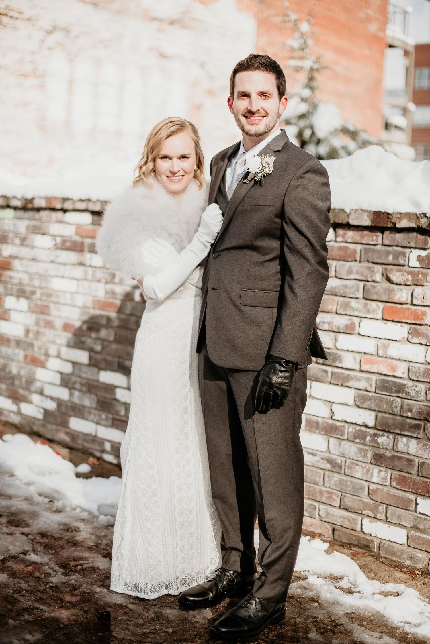 Bride in white lace gown with fur wrap and groom in gray suit pose together outdoors in winter snow by brick wall.