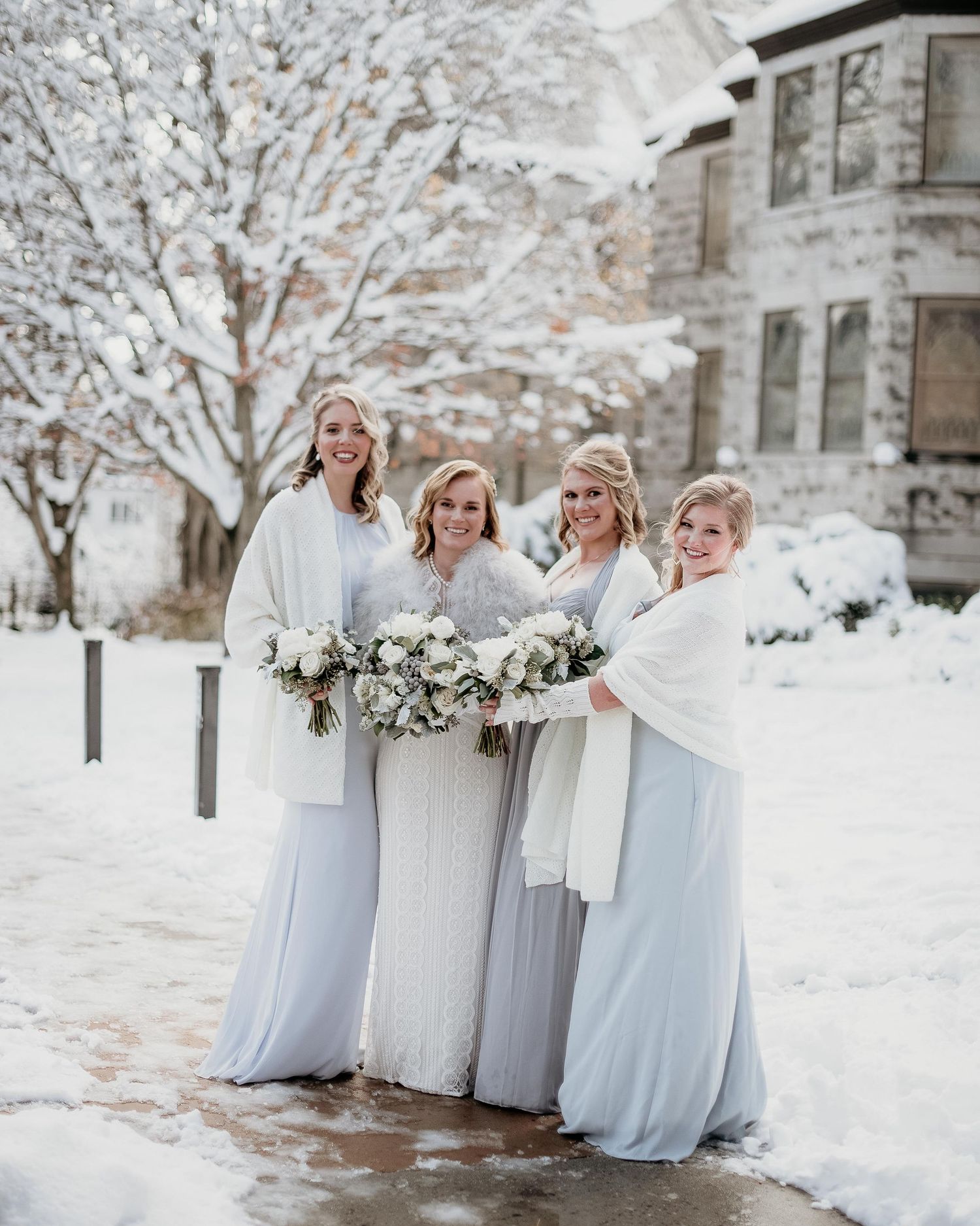 Bride and three bridesmaids in white and light blue gowns with wraps holding bouquets outdoors in a snowy winter setting.