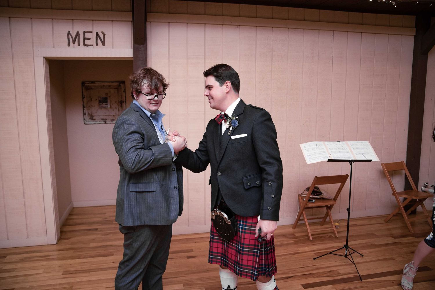 Two men conversing indoors, one wearing a traditional Scottish kilt with tartan pattern at a formal event.