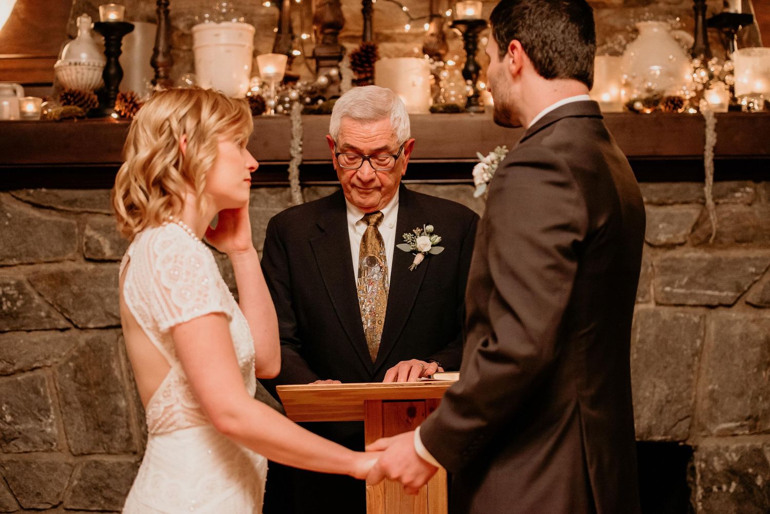 Bride and groom hold hands during intimate wedding ceremony officiated by elderly man before stone fireplace.