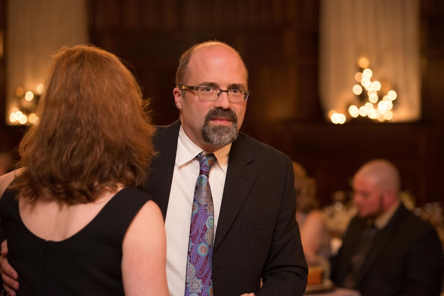 Man in suit and tie conversing with woman at formal indoor event with warm ambient lighting.