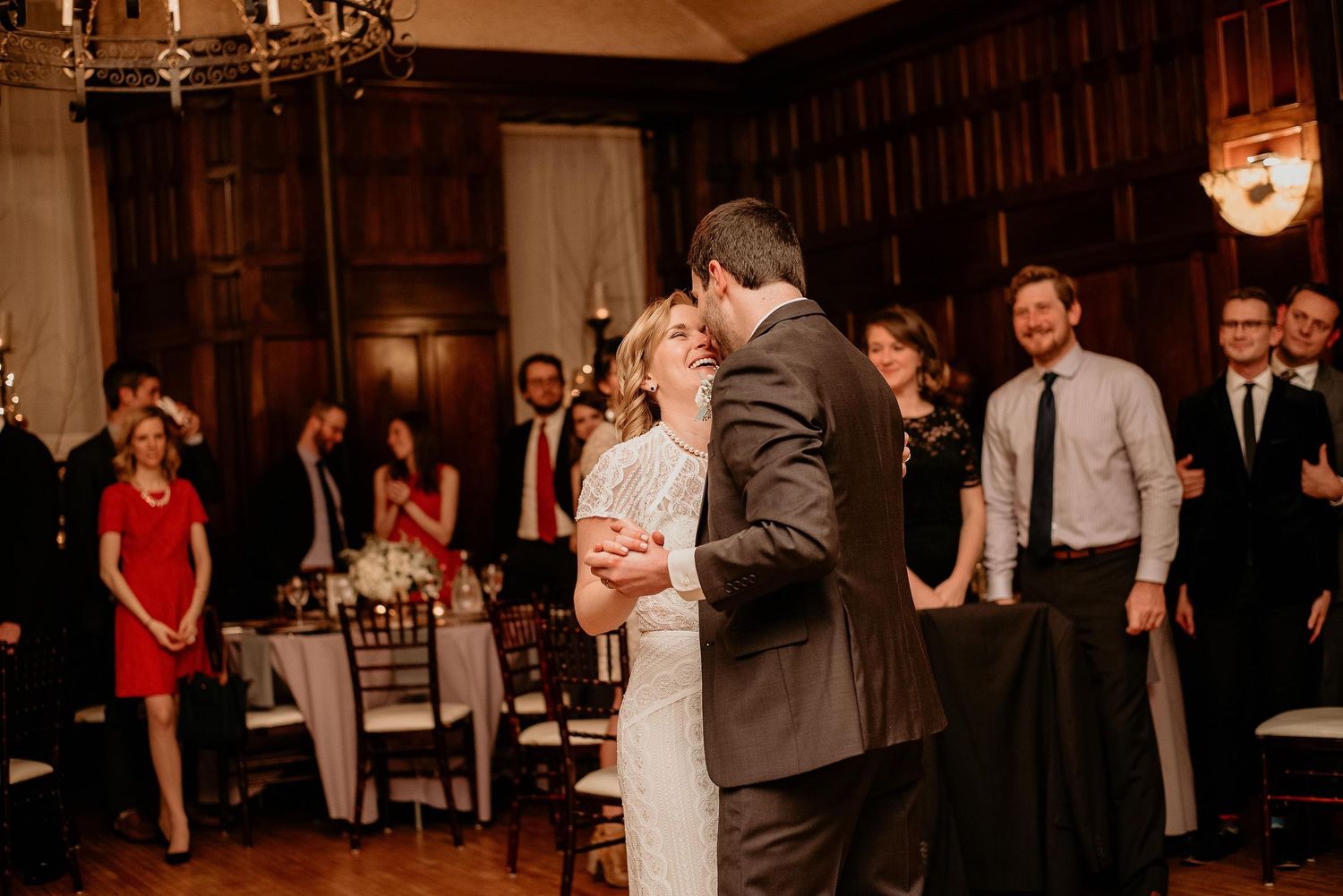 Bride and groom share first dance at elegant wood-paneled wedding reception while guests watch.