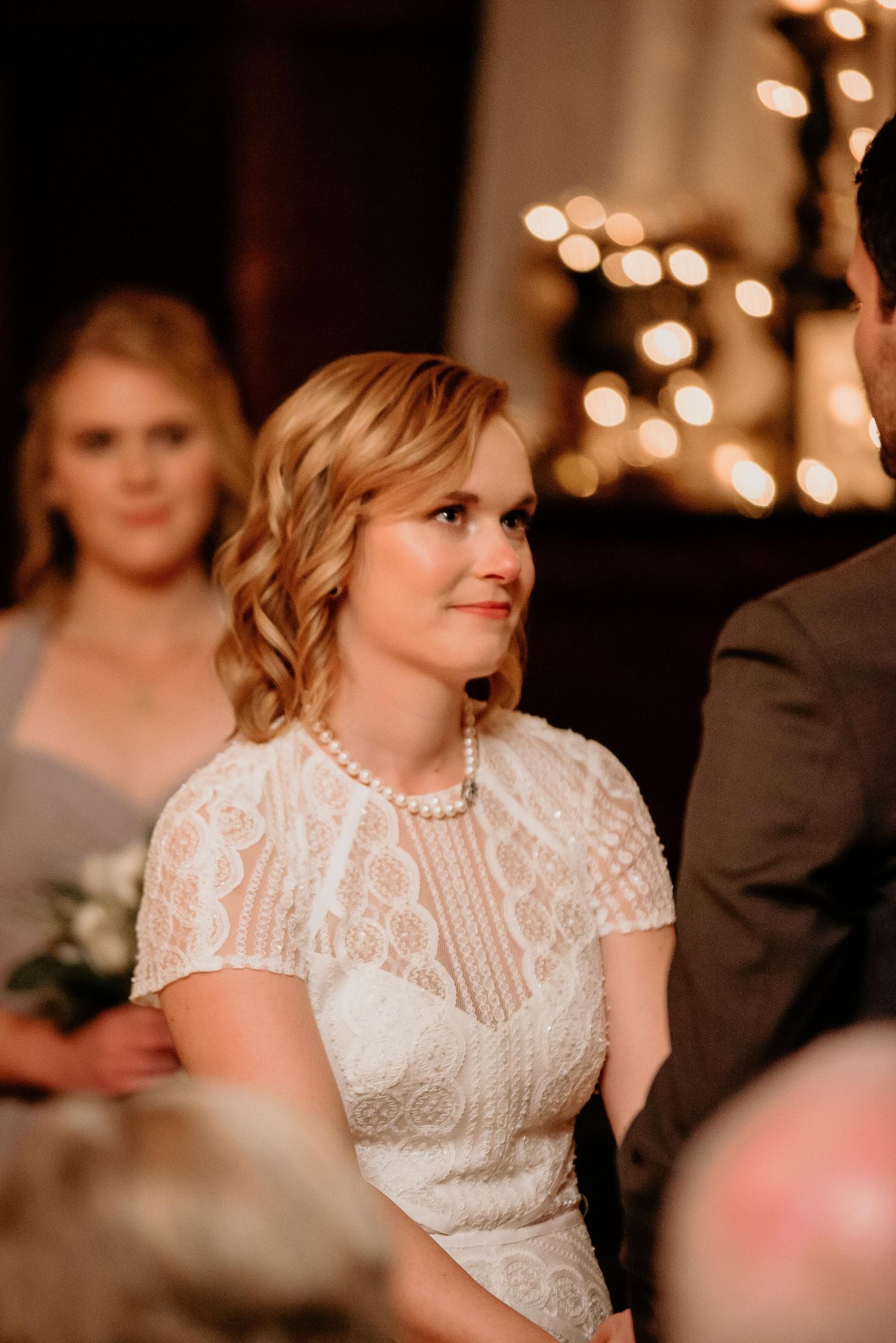 Bride in white lace dress and pearl necklace smiling during wedding ceremony with bokeh lights in background.