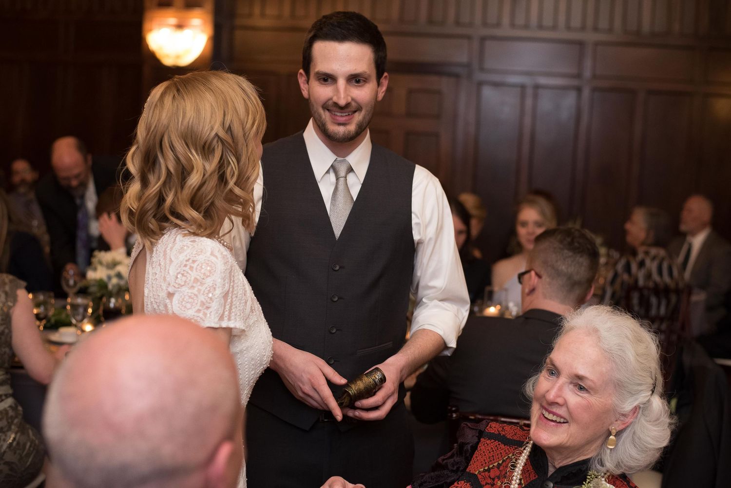 A groom in a black vest smiles at guests during an elegant indoor wedding reception in a wood-paneled venue.