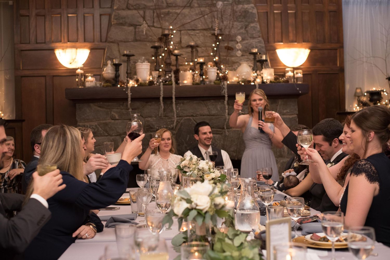 Wedding guests raising glasses in a toast at an elegant candlelit reception dinner table with floral centerpieces.