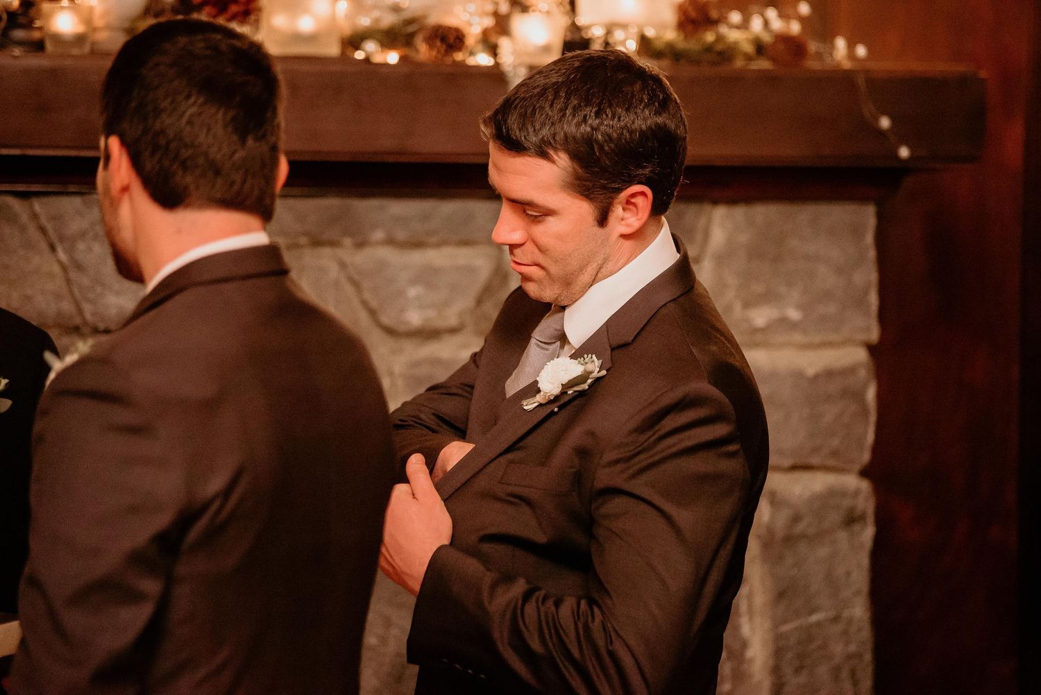 Two groomsmen in brown suits stand near a stone fireplace during a wedding reception.