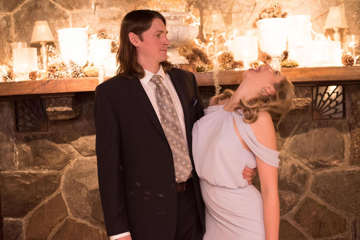 A couple in formal attire poses by a stone fireplace adorned with candles and pine cones at an elegant event.