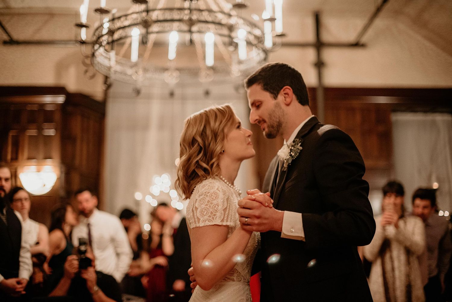 Couple shares first dance under chandelier at elegant wedding reception with guests watching in background.