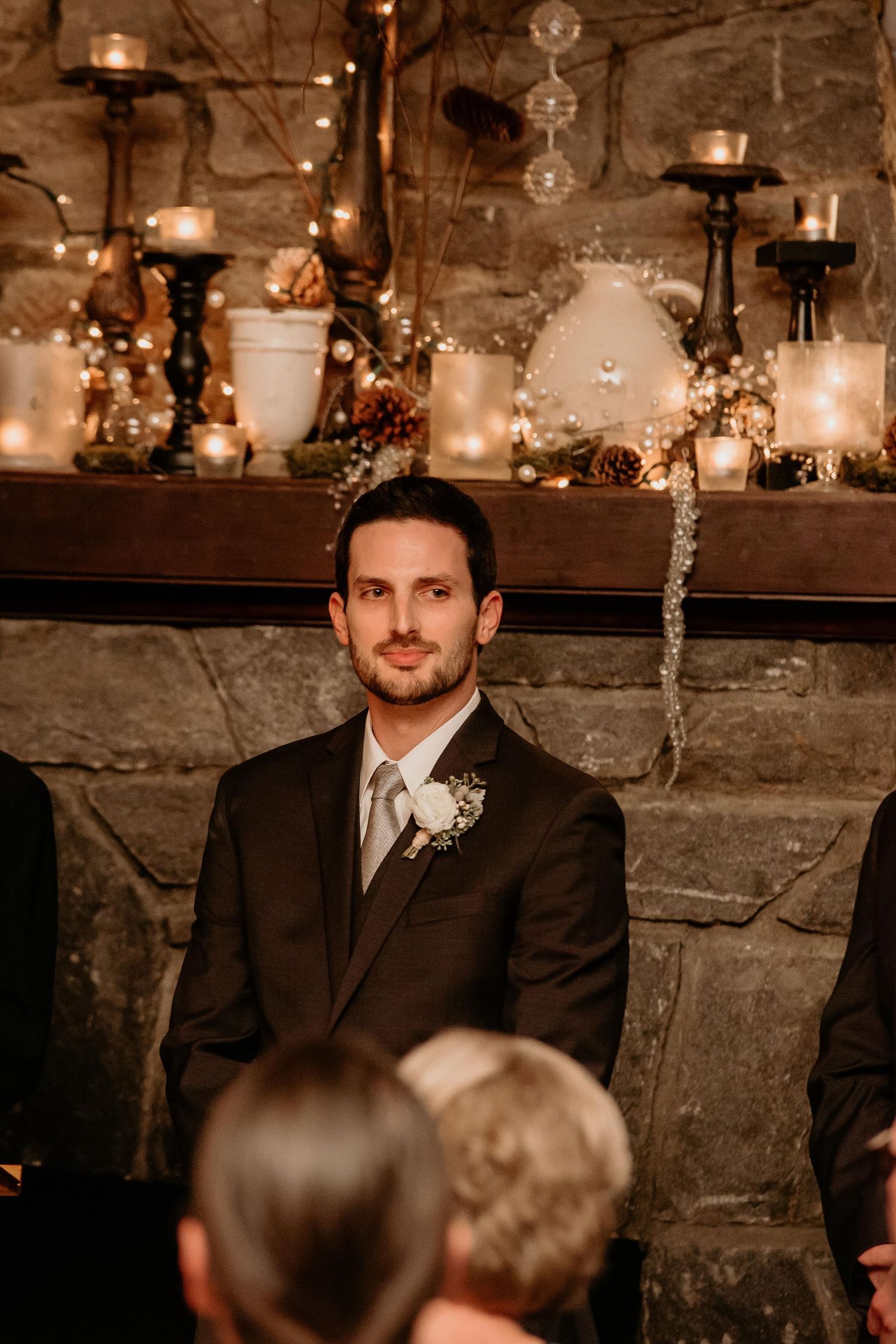 Groom in black suit with boutonniere stands before candlelit stone fireplace at elegant wedding ceremony.