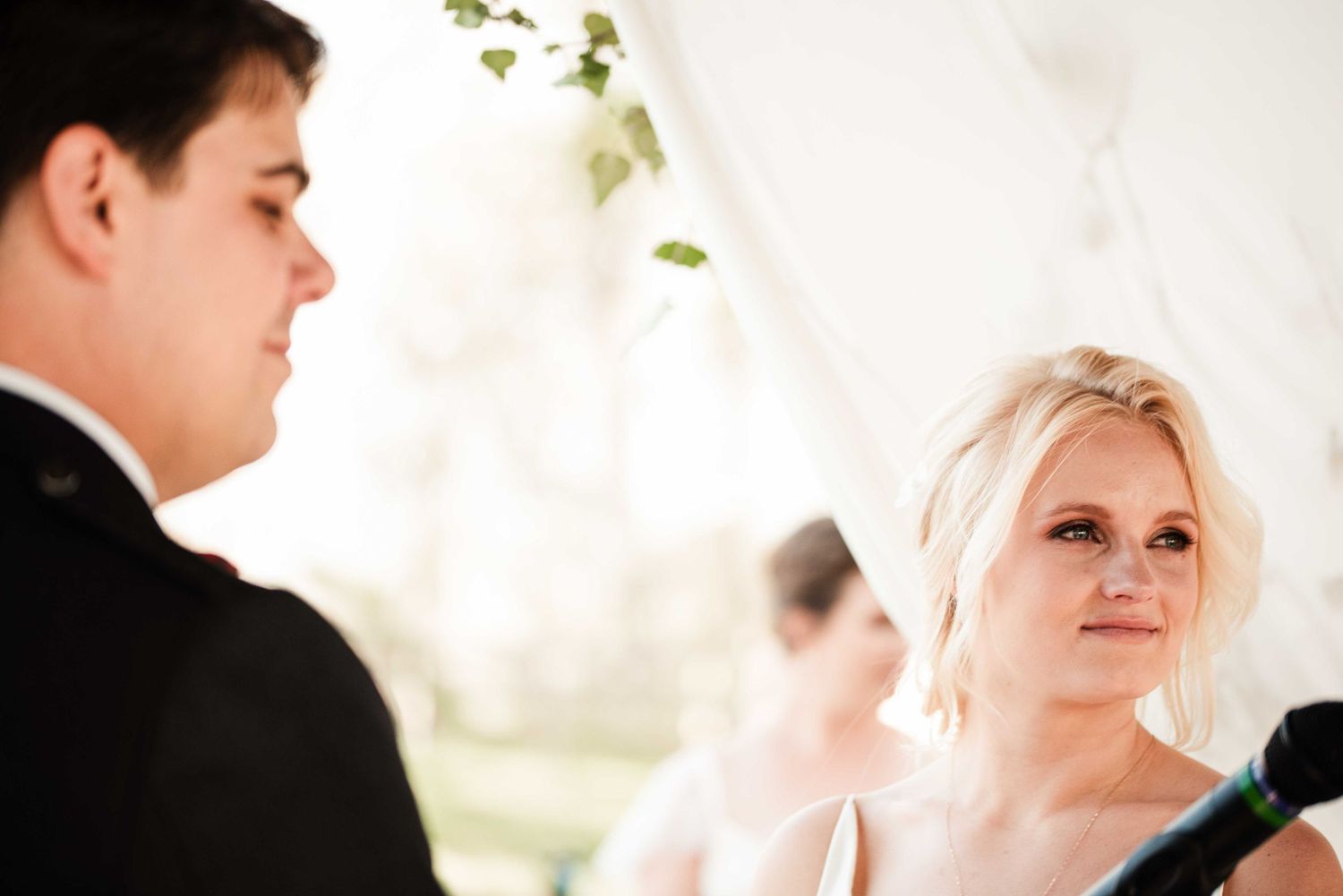 Bride gazing lovingly at groom during outdoor wedding ceremony under white tent.