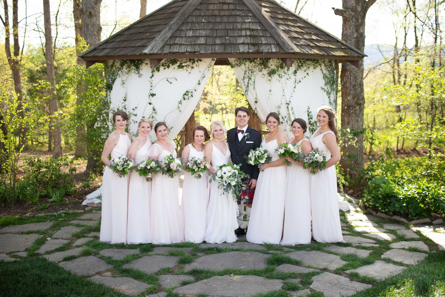 Bridal party in blush gowns and groom in kilt pose under floral gazebo arch outdoors in spring garden setting.