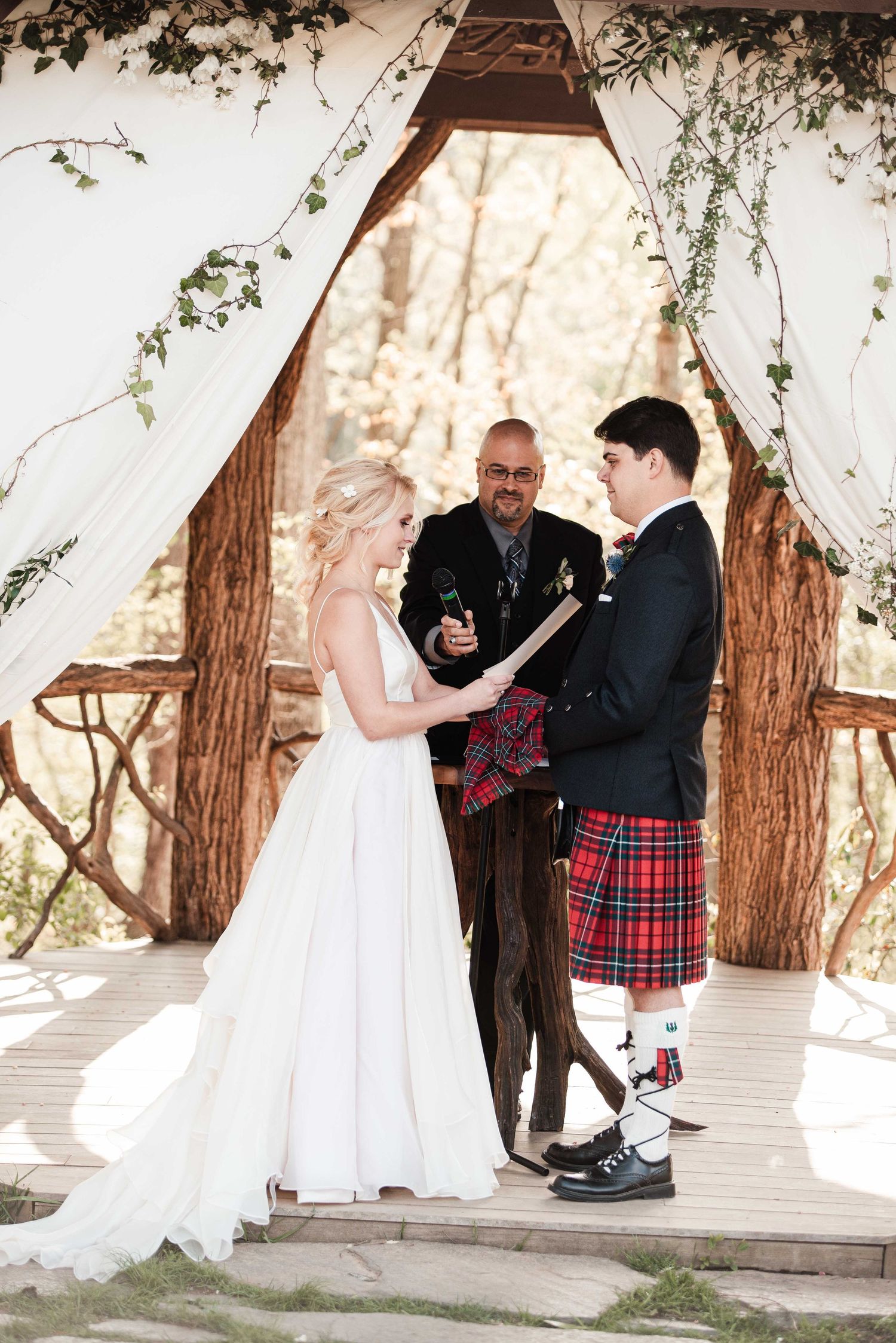 Bride and groom exchange vows under rustic wooden arch with white drapes, groom wearing red tartan kilt at outdoor wedding ceremony.