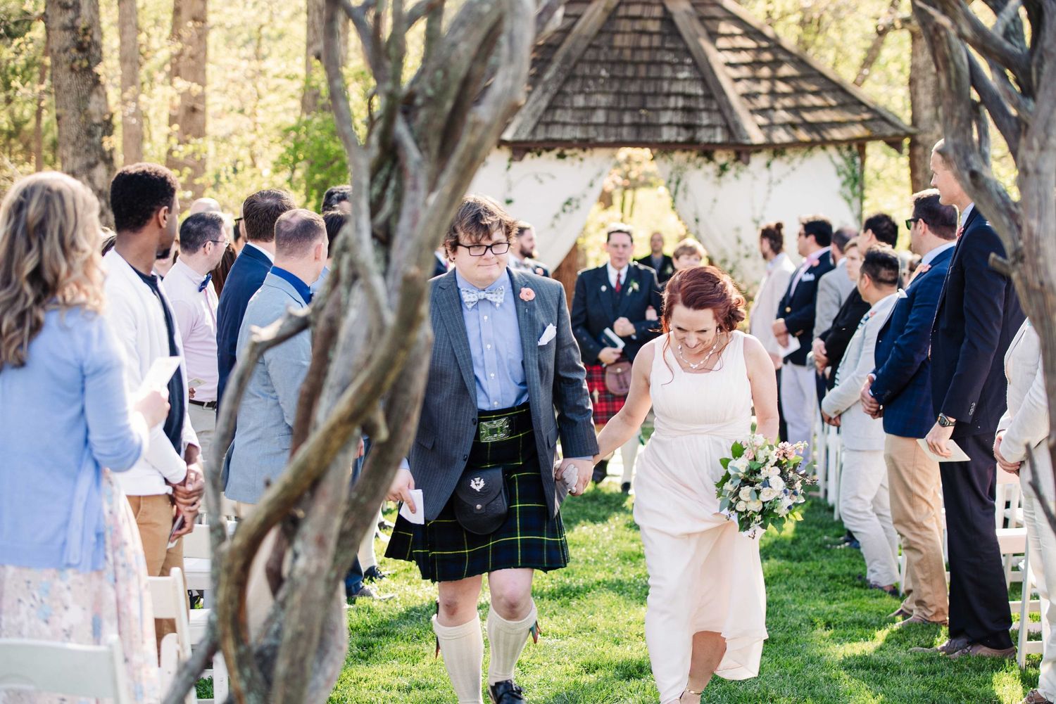 Bride and groom walk down outdoor wedding aisle, groom wearing tartan kilt, surrounded by seated guests in a garden setting.