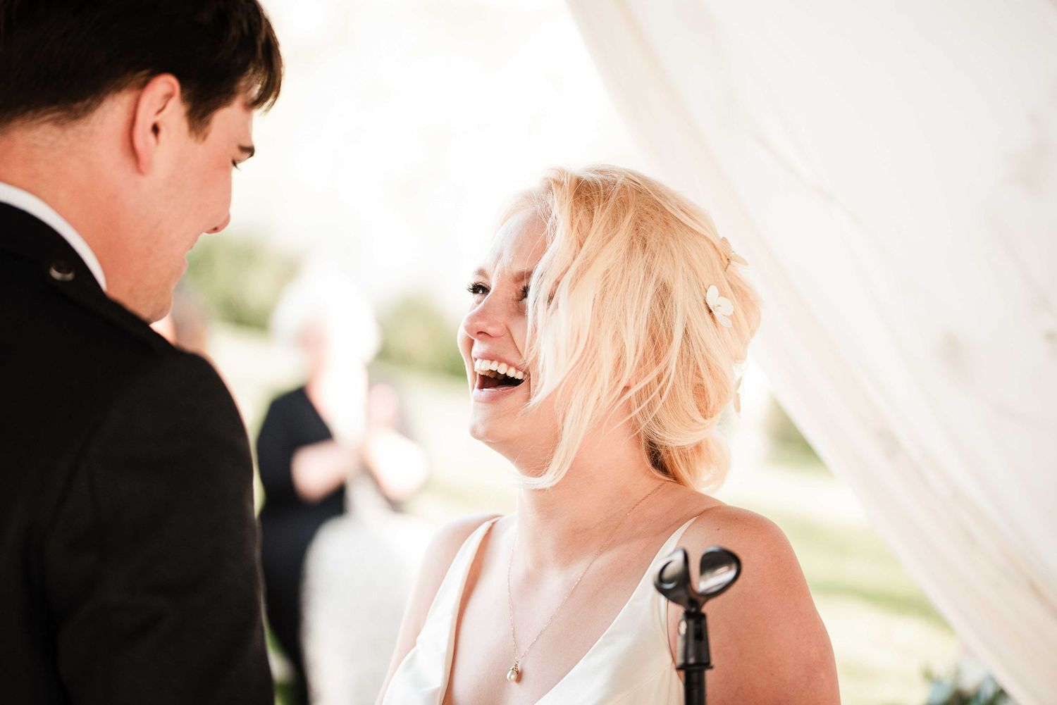 Bride laughing joyfully with groom during outdoor wedding ceremony under white draped tent.
