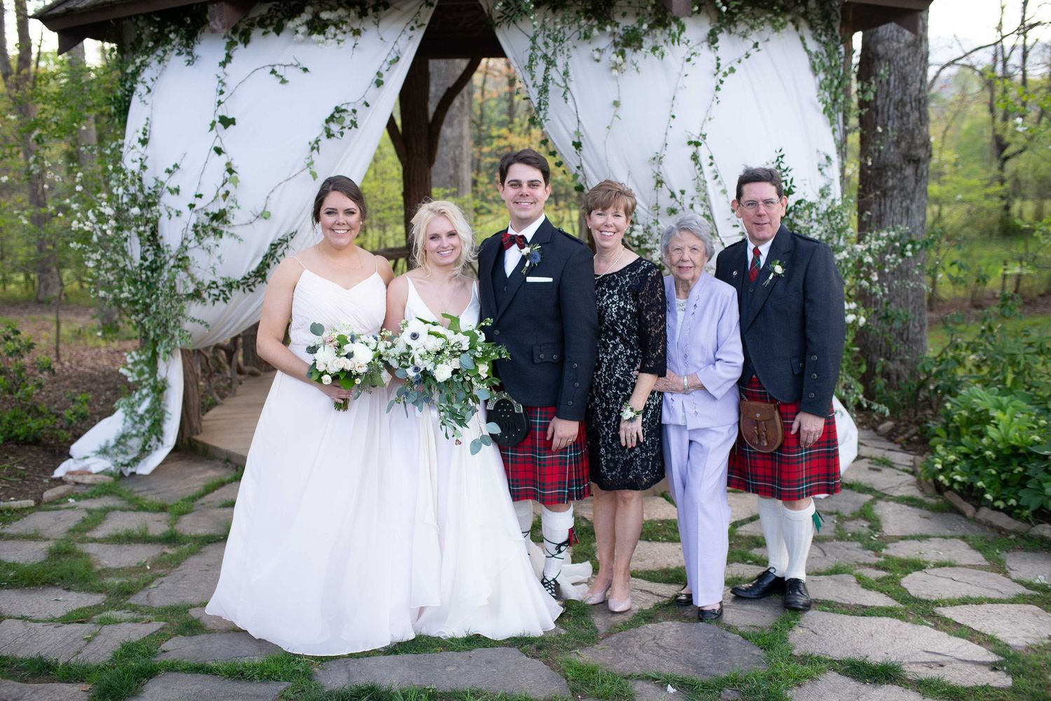 Wedding party posing under floral arch, groom and man in red kilts with bride, two women in formal attire.