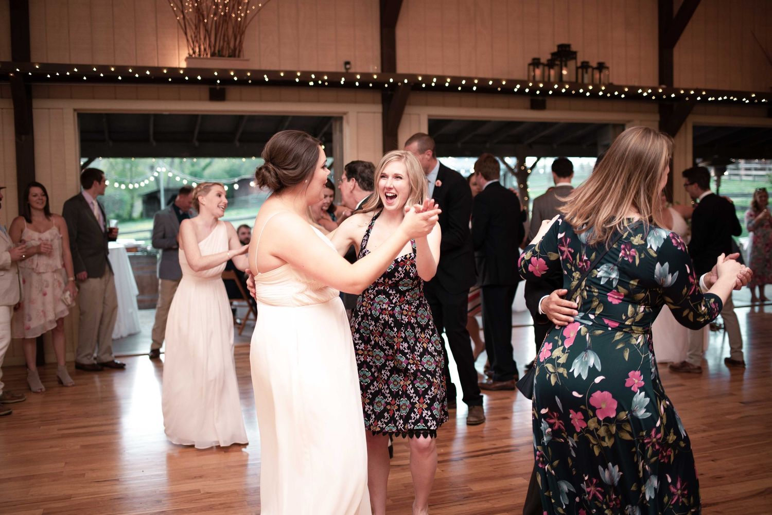 Wedding guests dancing together on the dance floor at a barn reception, including a woman in a white bridal gown.