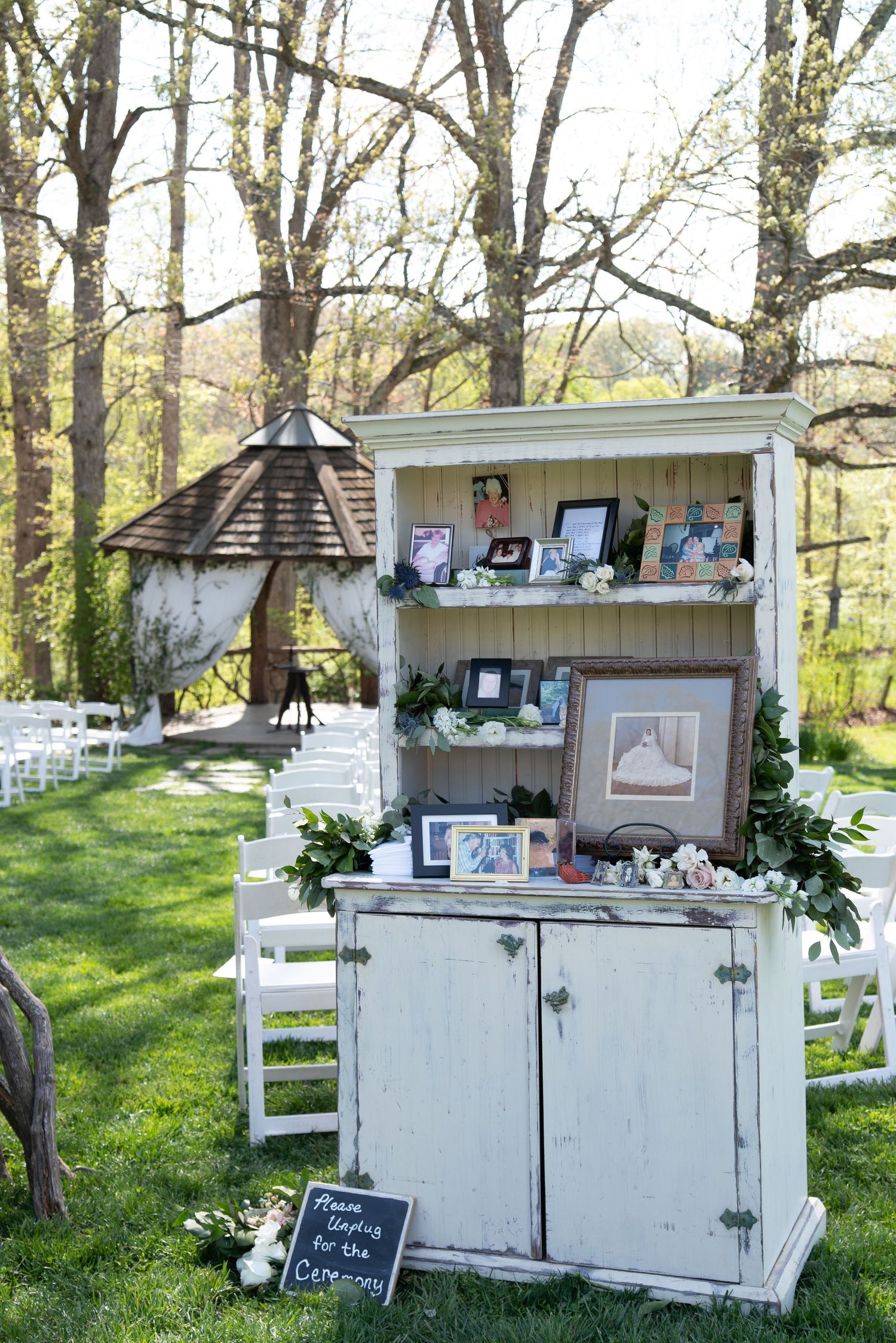 Rustic white cabinet memorial display with framed photos and greenery at outdoor wedding.