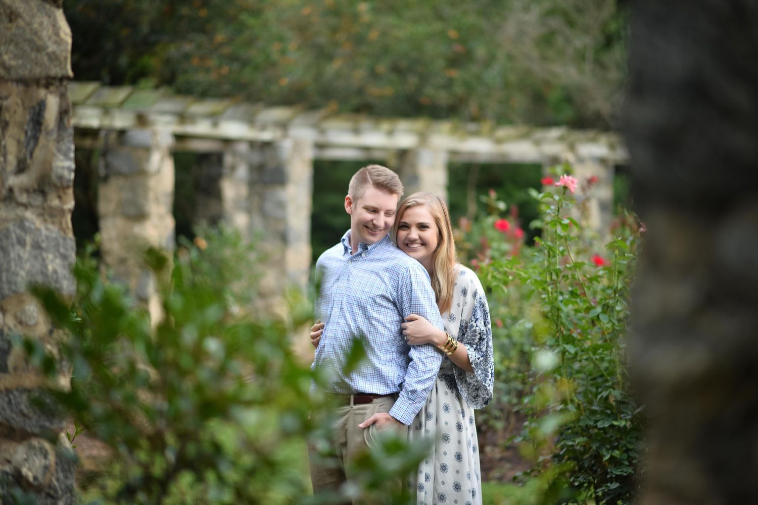 Couple embracing in a lush garden with stone ruins and red roses in the background.