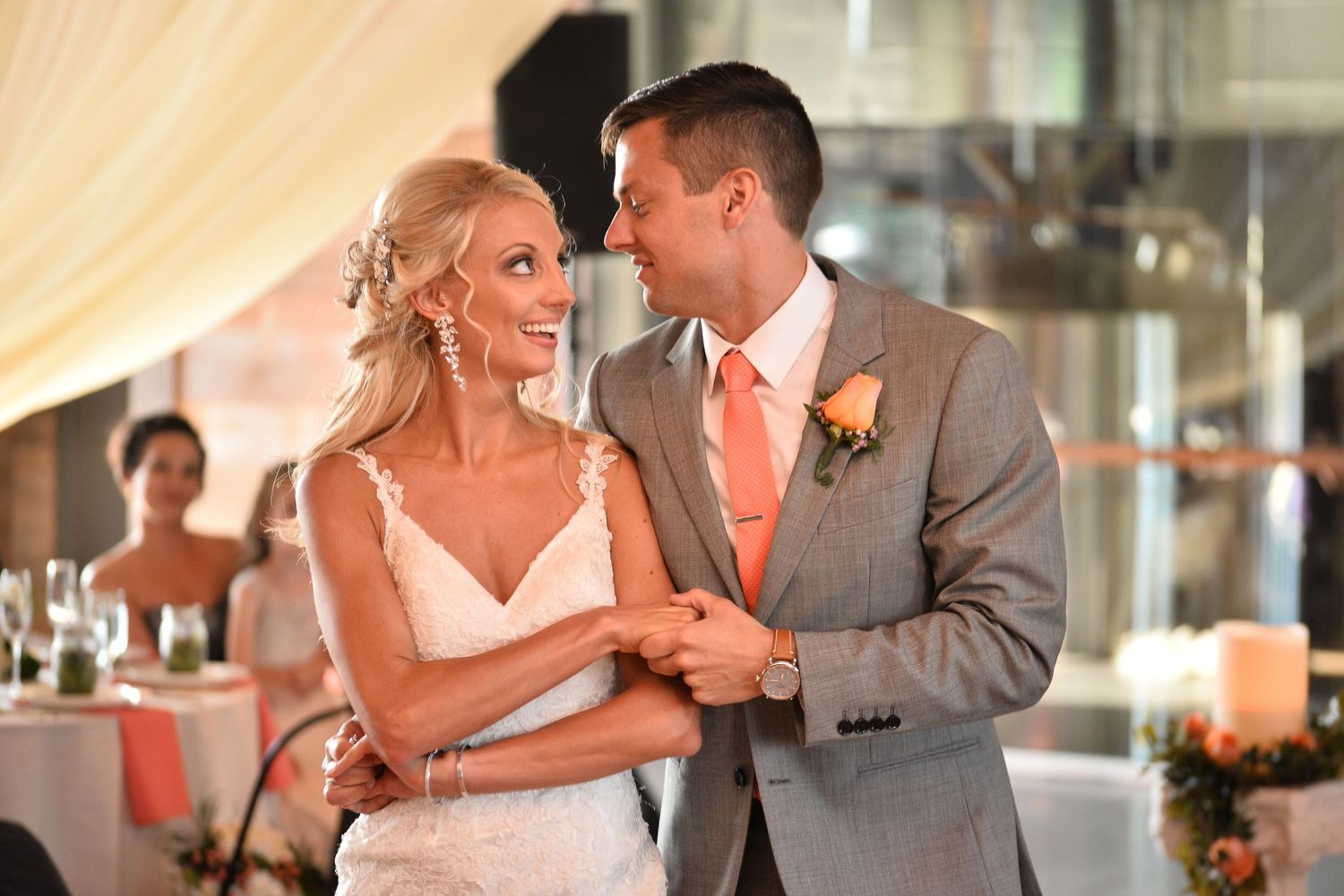 Smiling bride and groom in coral tie and gray suit share first dance at wedding reception.