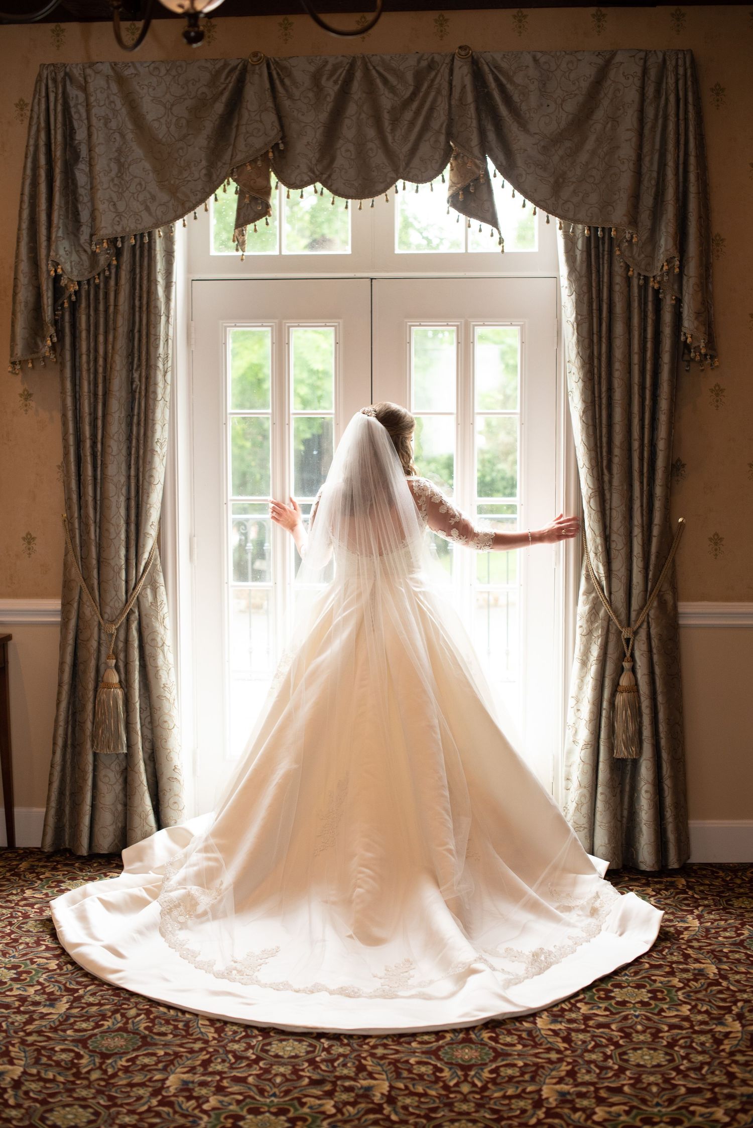 Bride in elegant ball gown with veil standing before large French doors in soft light.