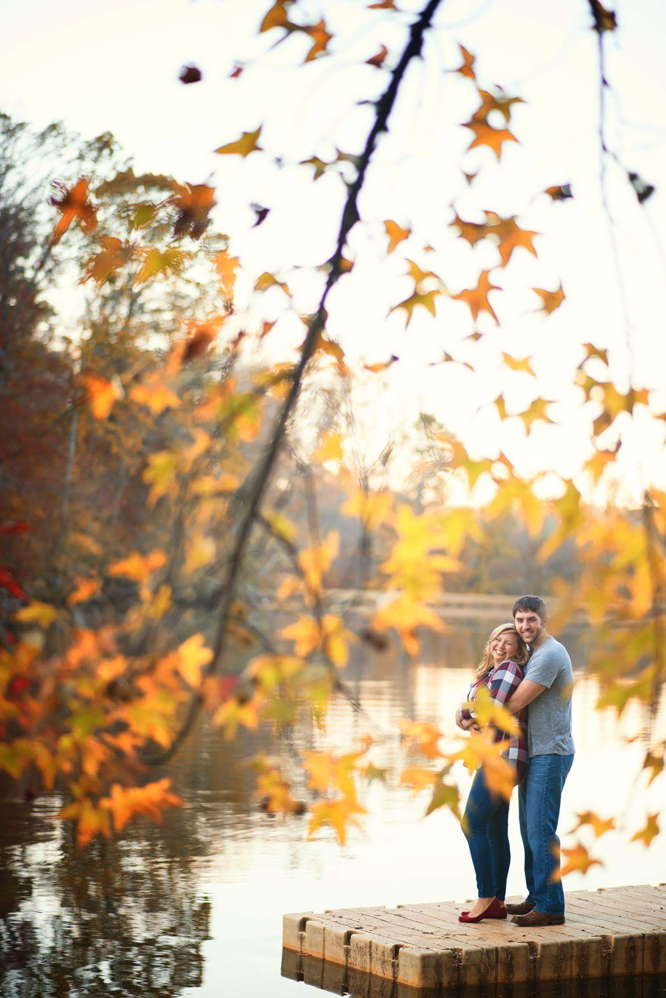 Couple embraces on dock surrounded by vibrant autumn foliage by lake.