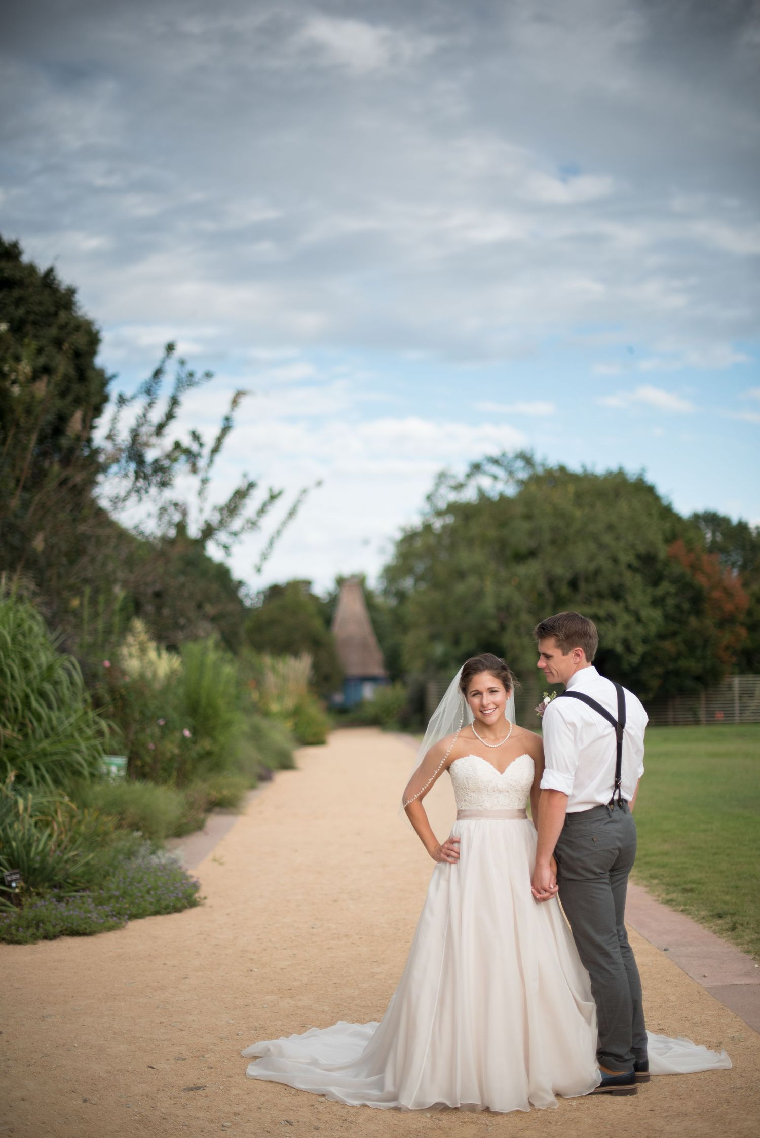Bride and groom holding hands on a garden pathway with lush greenery and cloudy sky backdrop.