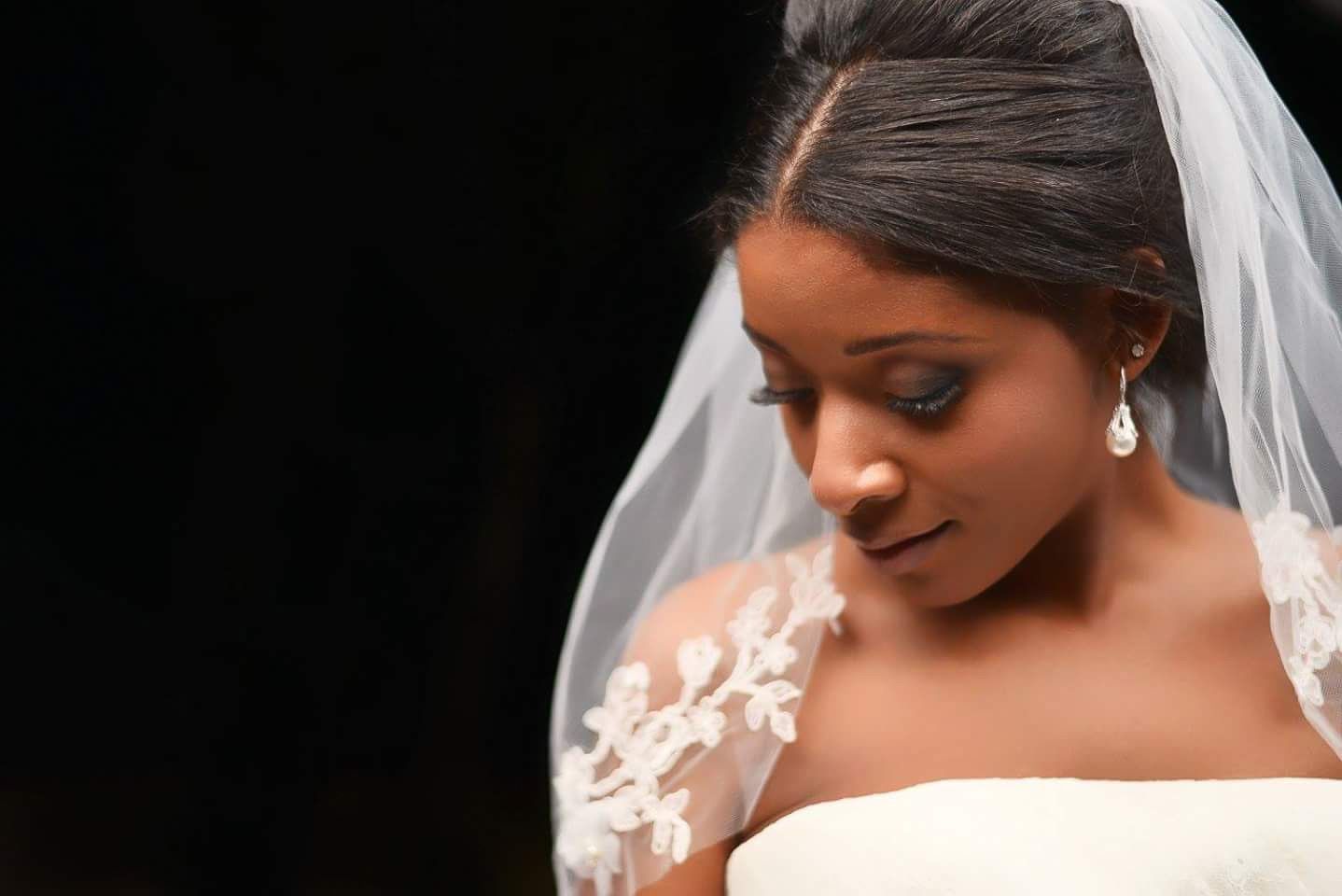 Beautiful African American bride in strapless gown with lace veil and pearl earrings gazes downward.