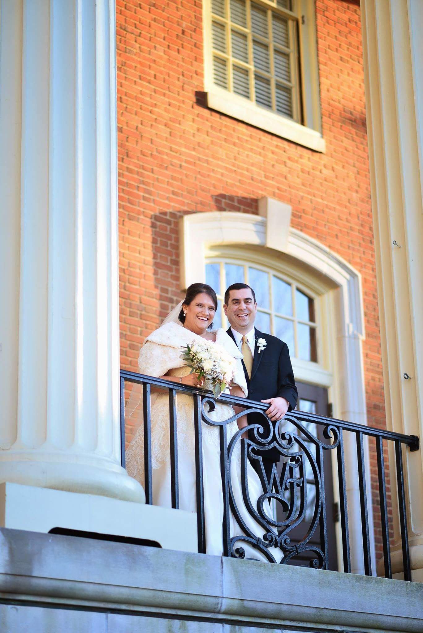 Smiling bride and groom posing on elegant brick building balcony with ornate iron railing.
