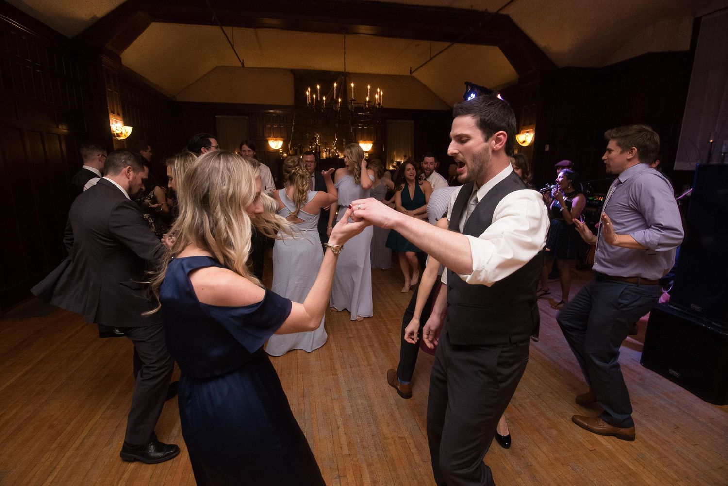 Guests dancing joyfully at an elegant wedding reception in a warmly lit ballroom with chandeliers.