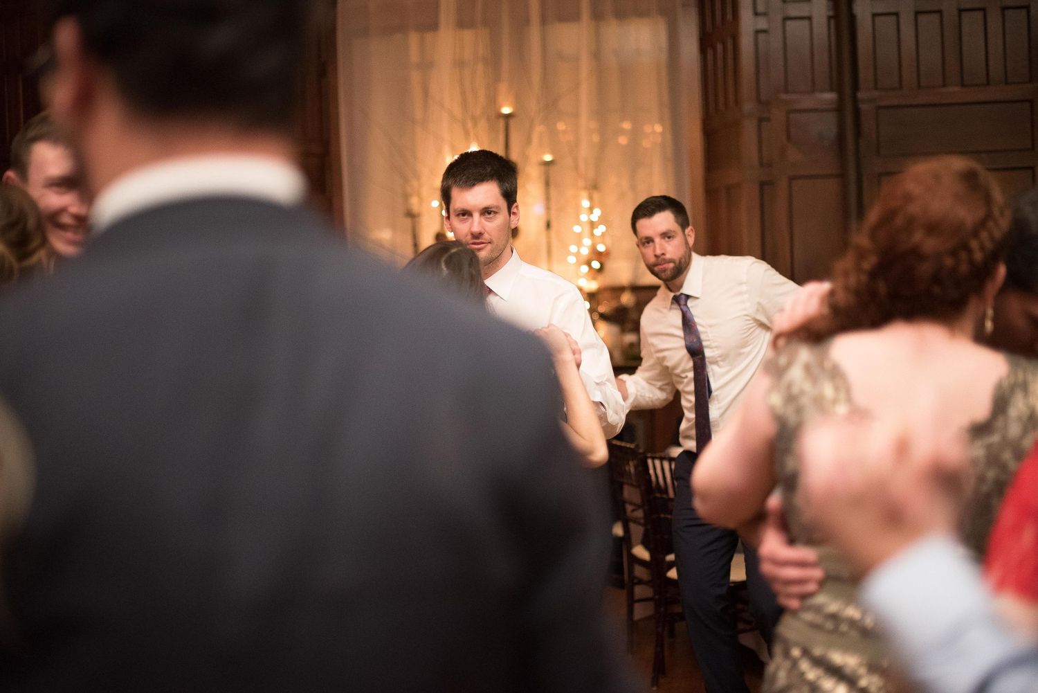 Two men in formal attire watch guests dance at an elegant candlelit wedding reception.
