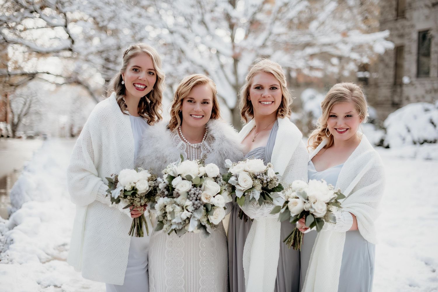 Four bridesmaids in white and gray gowns holding white floral bouquets posing outdoors in a snowy winter wedding scene.