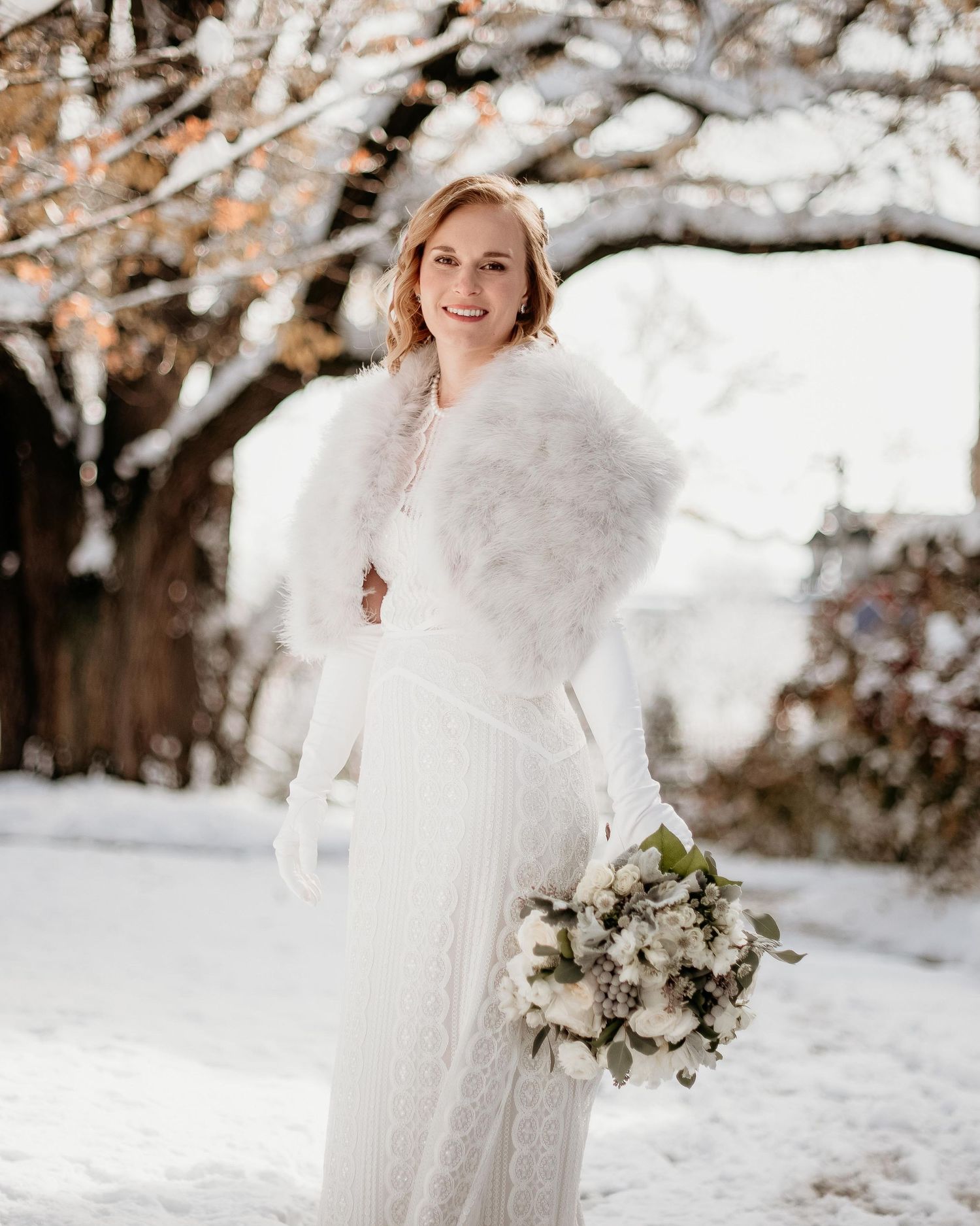 Bride in white lace gown and fur stole holds bouquet in snowy winter setting.