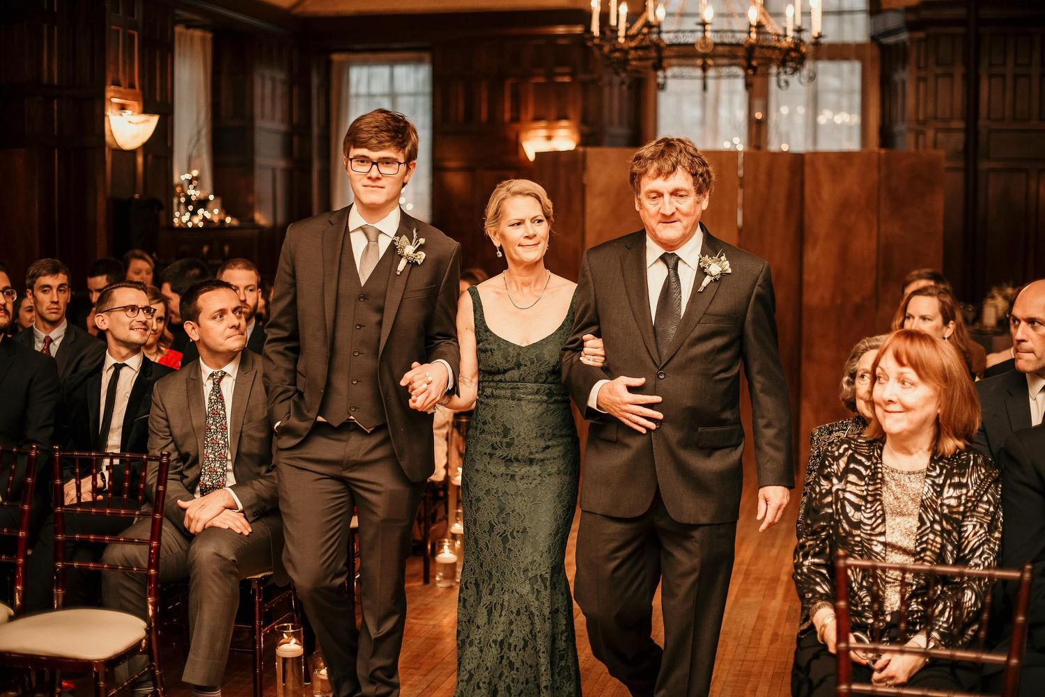 A woman in a green gown walks down the aisle escorted by two men in suits at an elegant wood-paneled wedding ceremony.