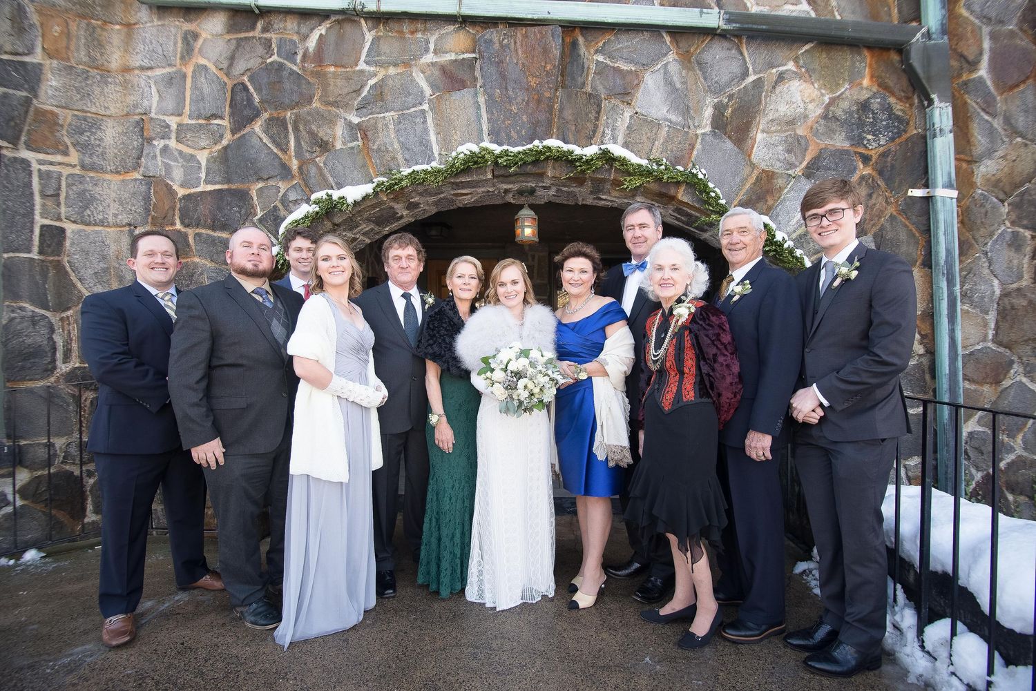 Wedding party posing in front of a stone church entrance in winter, bride in white gown with family and attendants.