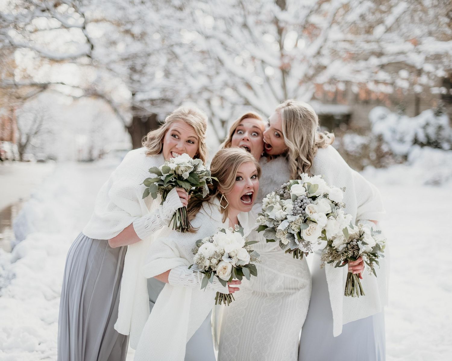 Bride and bridesmaids in white dresses holding floral bouquets laughing together in a snowy winter wedding outdoor setting.