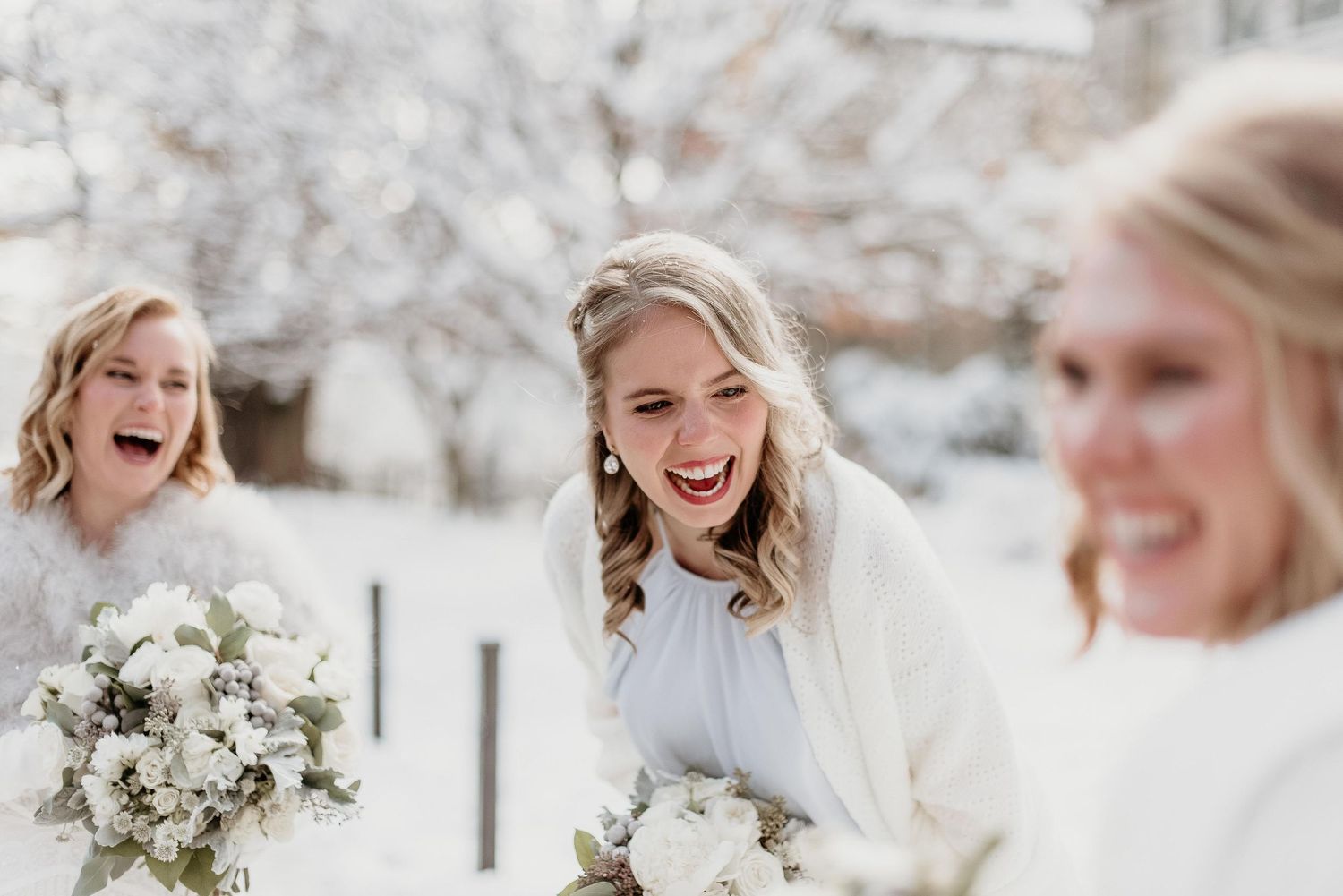 Bride and bridesmaids laughing outdoors in winter snow, holding white floral bouquets in elegant white attire.