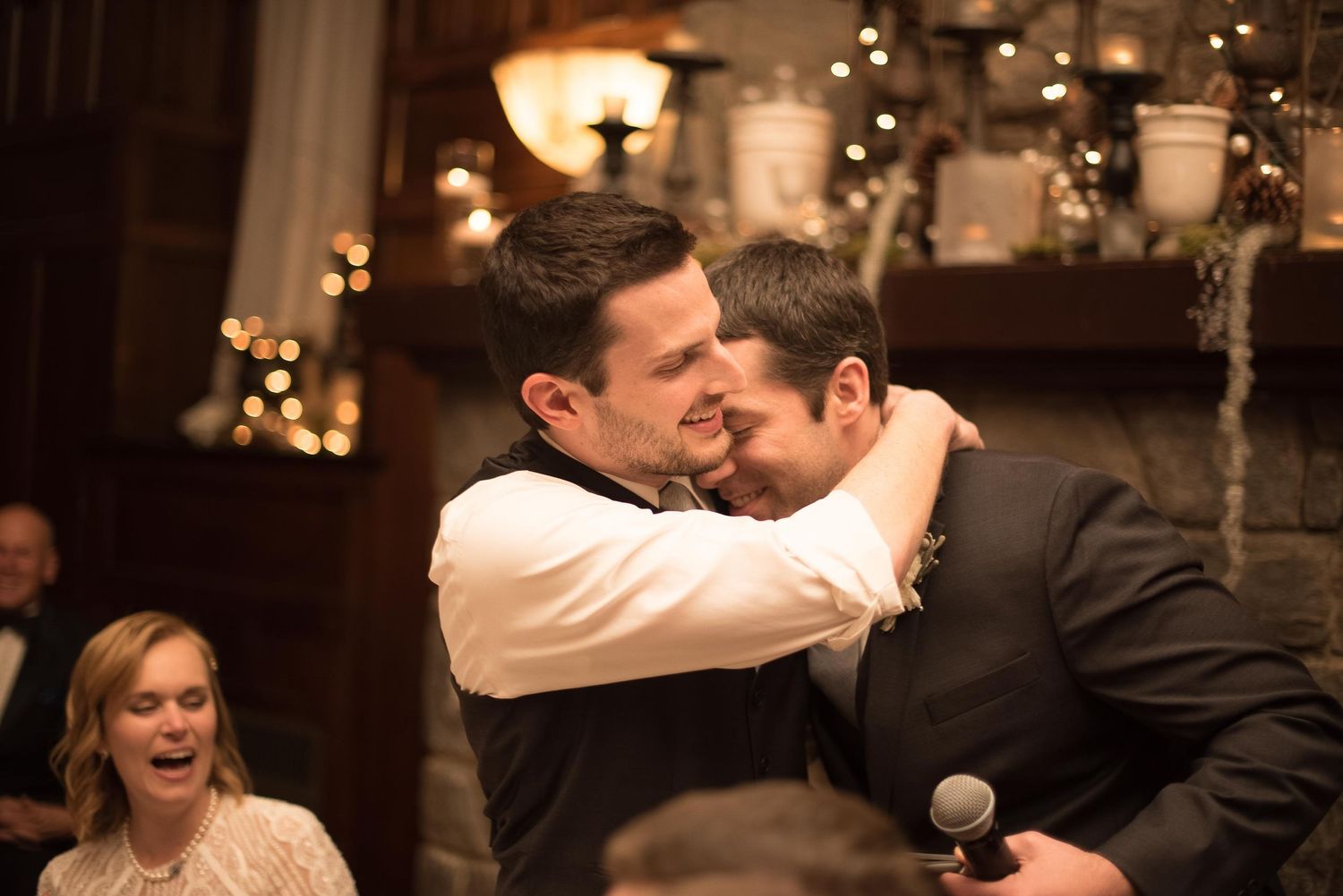 Two men share an emotional hug at a wedding reception while guests smile and laugh in the background.