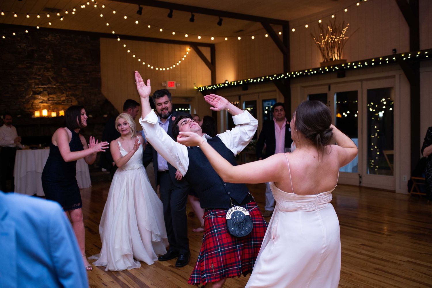 Guests dancing joyfully at a wedding reception in a rustic venue with string lights and a man in a kilt.