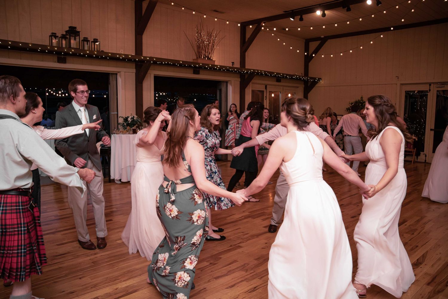 Guests dancing in a circle at a wedding reception in a rustic barn venue with string lights.