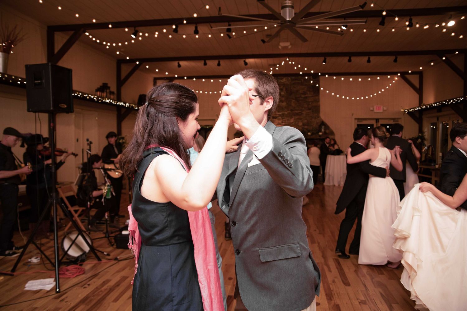Couple dancing together at a rustic wedding reception venue with string lights and live band in background.