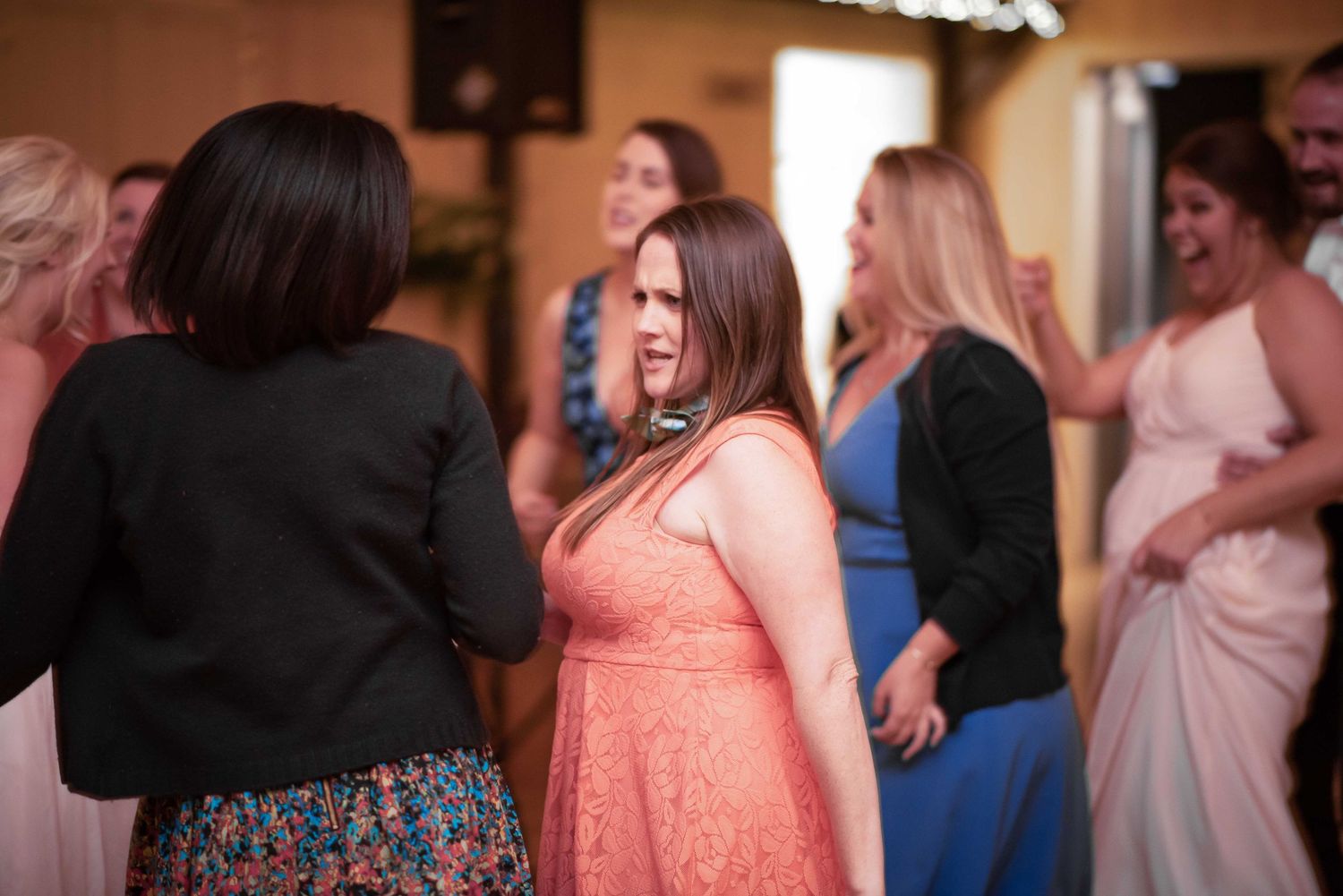 Women in colorful dresses dancing and socializing at an indoor wedding reception celebration.