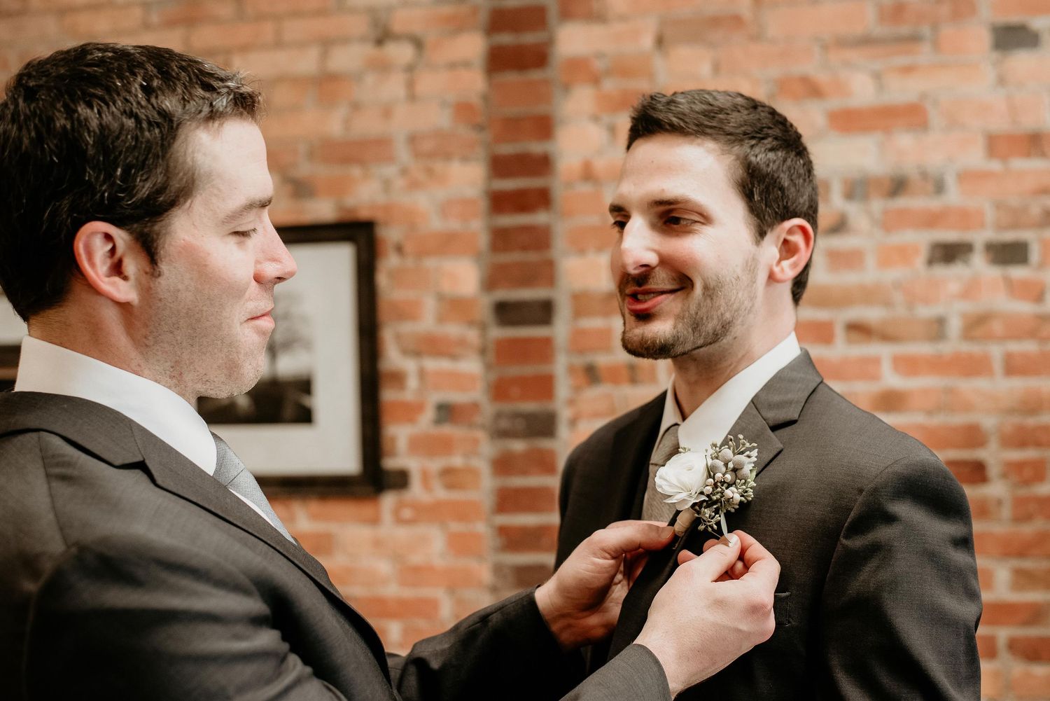 A groomsman pins a boutonniere on the groom's lapel in front of a rustic brick wall.
