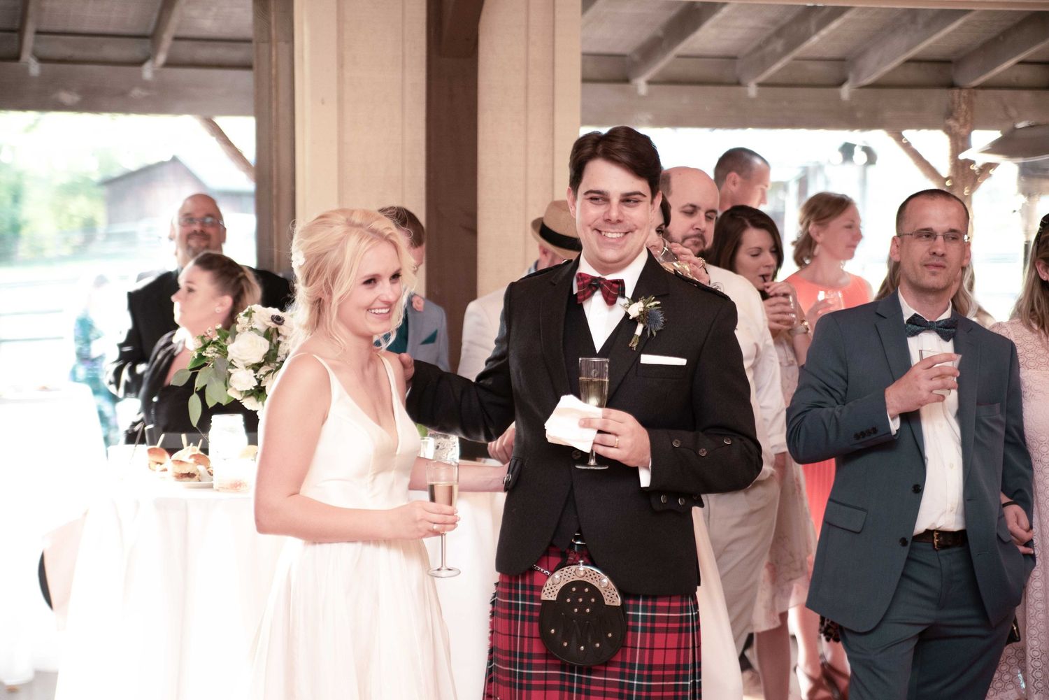 Bride in white gown and groom in red tartan kilt smile holding drinks at elegant wedding reception.